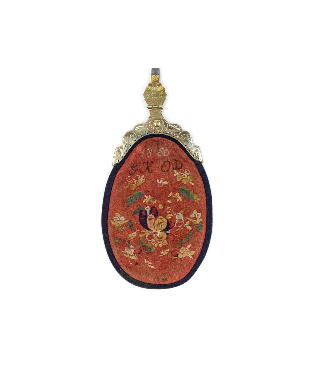 Brodert lomme med messinglås. Broderi med floralmønster, årstallet "1850" og initialer "GKOD". Messinglås gravert med ranker og initialene "ROD". To stykker løst originalt fór - én av skinn og én av lin.