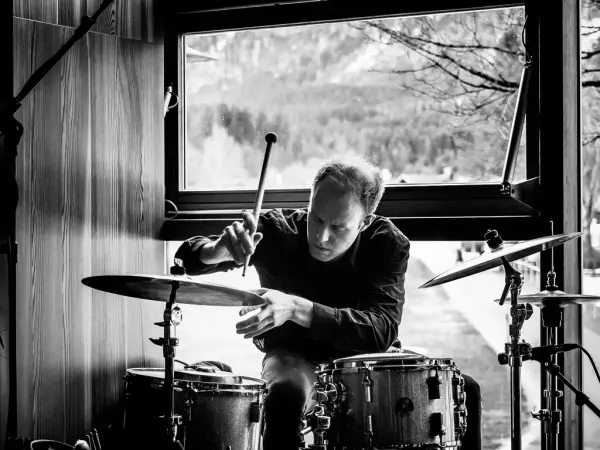 Her ser vi en mann som sitter bak et trommesett. Han sitter foran et vindu med obskur natur i bakgrunnen. Bildet er svart hvitt.