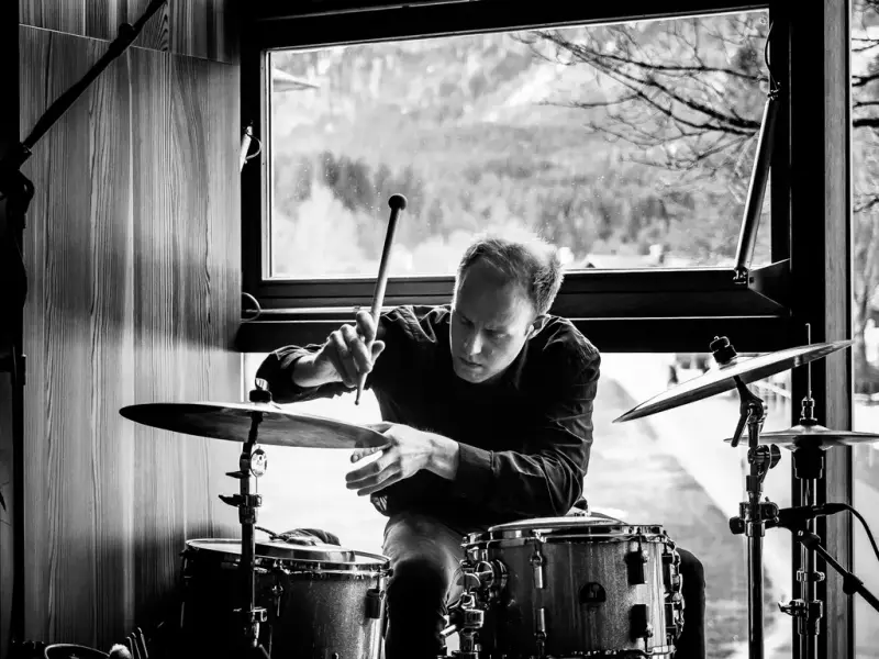 Her ser vi en mann som sitter bak et trommesett. Han sitter foran et vindu med obskur natur i bakgrunnen. Bildet er svart hvitt.