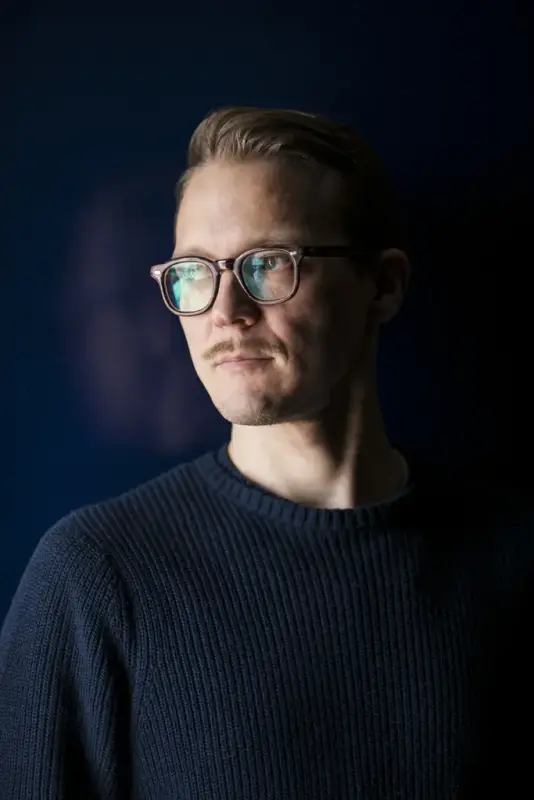 Profilfoto av mann med briller som ser ut mot siden. Hand har blå genser, brunt hår og står foran en blå bakgrunn. 