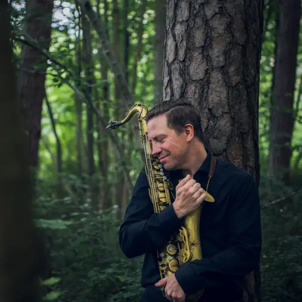 Bildet viser en man som omfavner en saksofon mens han smiler. Hans står lent inn mot et tre i en skog.