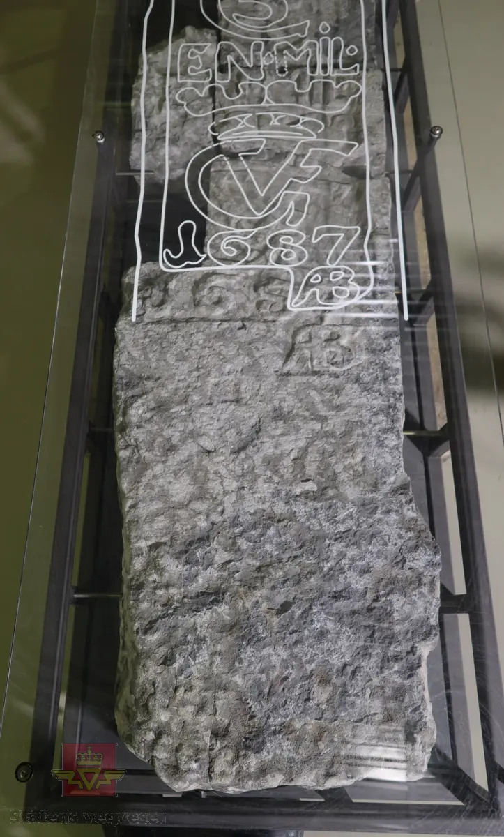 Grå kalkstein i 5 deler. Noe av monogrammet er borte, men 1687 er godt synelig. Kun den største delen er merket. Steinen er lagret på to paller.