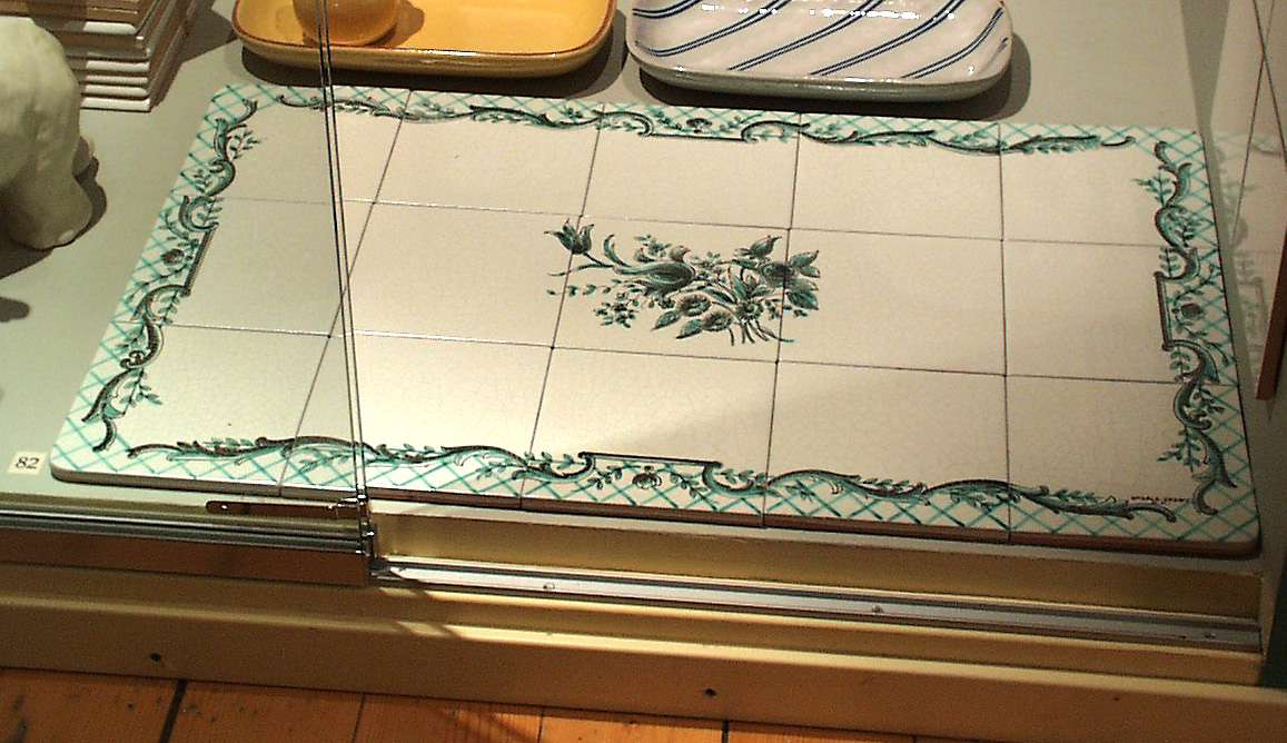 15 st kakelplattor till bordskiva, av gult lergods med målad dekor bestående av bladslingor och rutor i grönt och svart samt kantbård mot grå vit krackeléglasyr, de fyra hörnplattorna har rundade hörn. På baksidan i relief: 1045 8 UE.