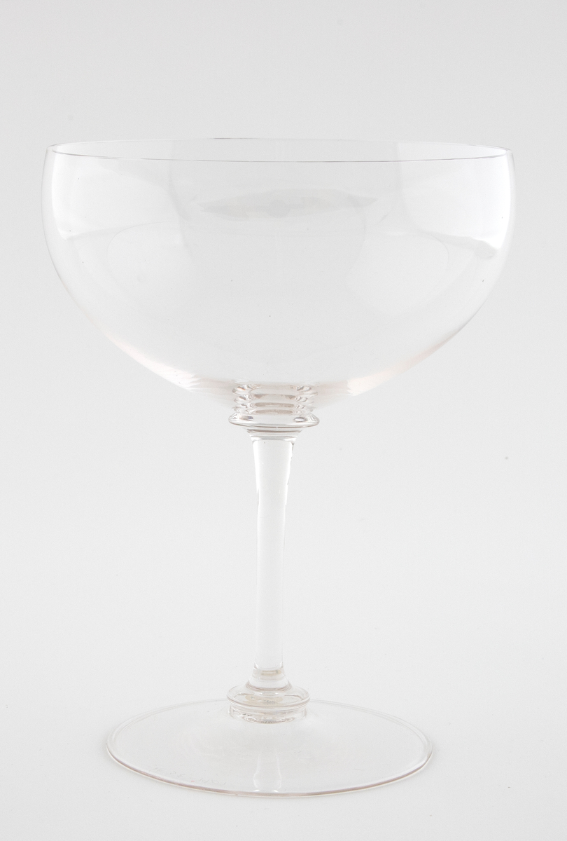 Drikkeglass i klart glass. Lav og vid kupa, som hviler på en smal stett. Sirkulær fotplate, noe hevet på midten. Overgangen mellom fot, stett og kupa er markert med ringformede vulster.
