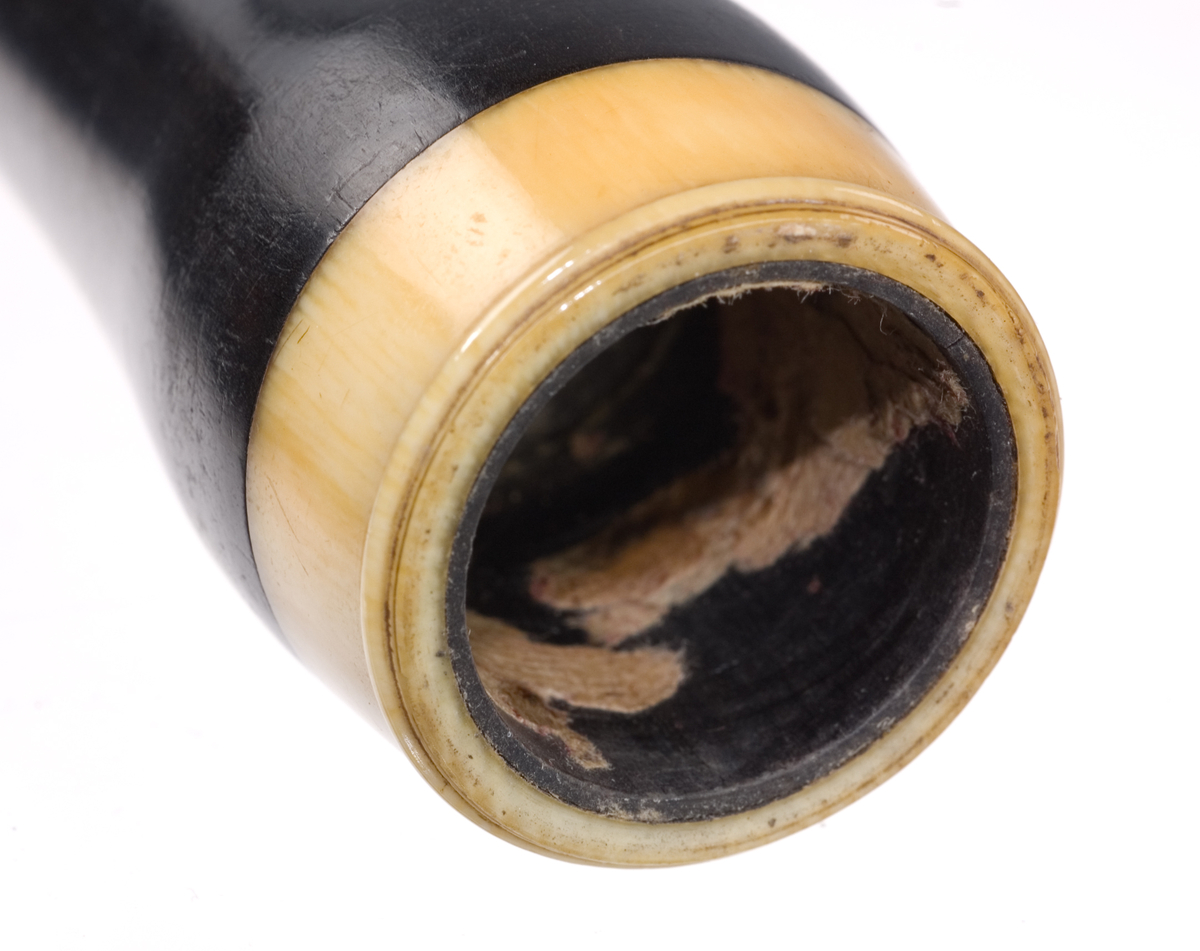 Tvärflöjt i ebenholts med ringar i elfenben. Kortare flöjt, troligtvis stämd i Eb eller F. 4 delar. Ovalt anblåsningshål. 2 klaffar, den övre med bocklagring, den lägre med vulstlagrin. Kvadratiska plattor.