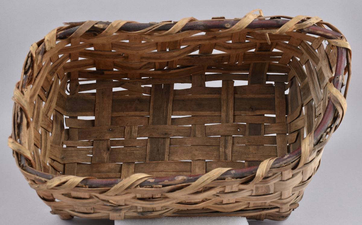 Korg i rakflätad spån. Kantens vidja är lindad. Rektangulär form med flätad botten.