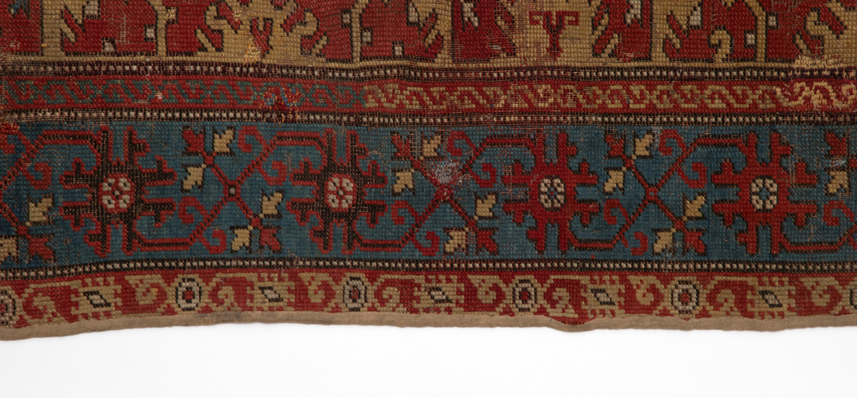 Håndlaget rektangulært teppe i ullfloss, utført med ghiordesknuter. Tett sammenhengende geometriske figurer i gult med litt blått og med svarte konturer på rød bunn. En klar blå bord rammer inn midtmotivet. Denne er dekorert med svarte og røde stiliserte blomster i ranke.