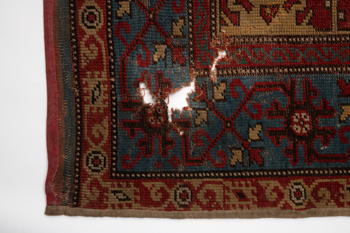 Håndlaget rektangulært teppe i ullfloss, utført med ghiordesknuter. Tett sammenhengende geometriske figurer i gult med litt blått og med svarte konturer på rød bunn. En klar blå bord rammer inn midtmotivet. Denne er dekorert med svarte og røde stiliserte blomster i ranke.