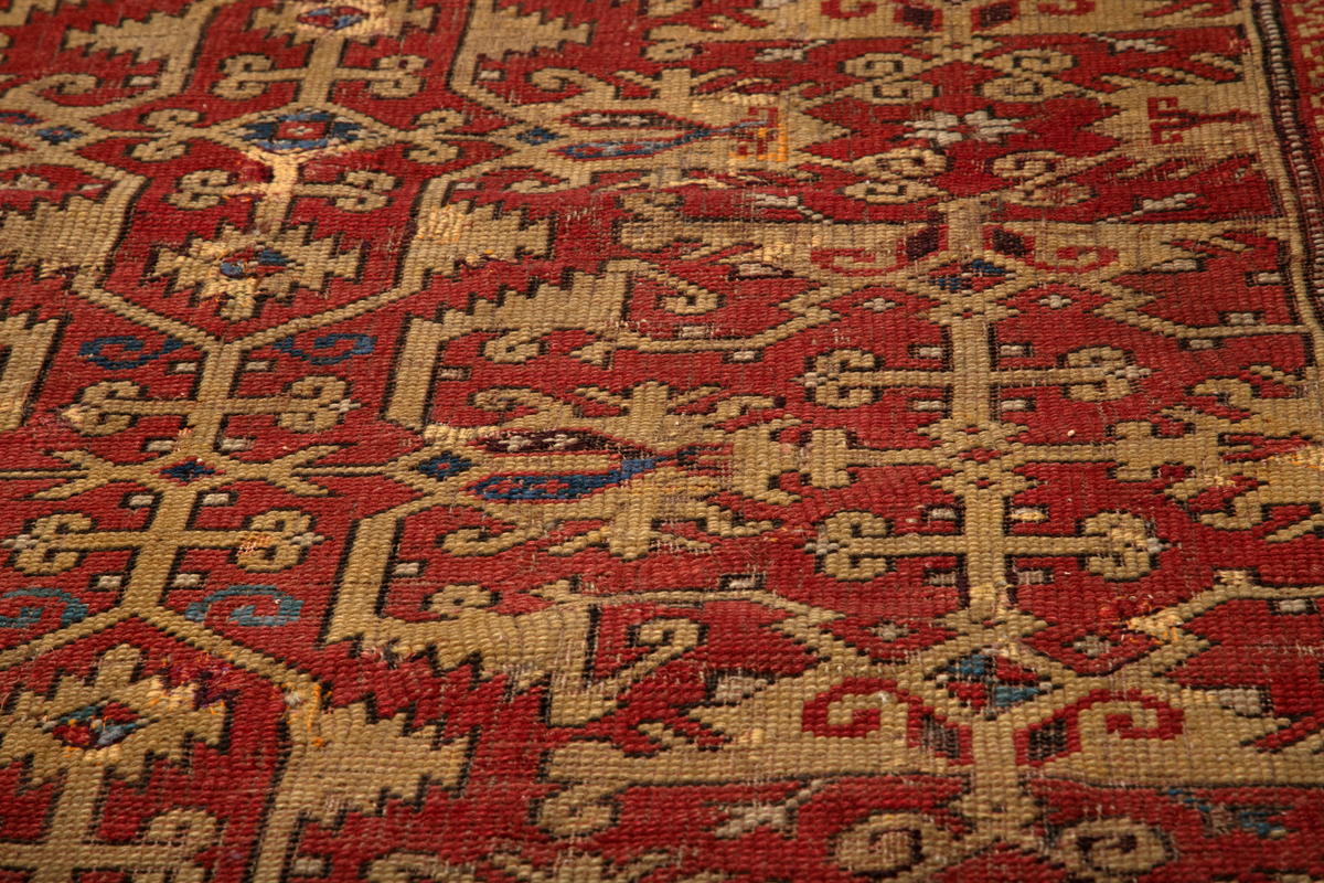 Håndlaget rektangulært teppe i ullfloss, utført med ghiordesknuter. Tett sammenhengende geometriske figurer i gult med litt blått og med svarte konturer på rød bunn. En klar blå bord rammer inn midtmotivet. Denne er dekorert med svarte og røde stiliserte blomster i ranke.