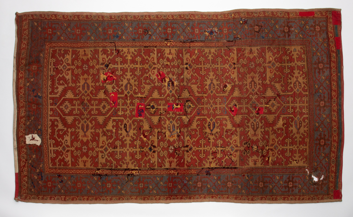 Håndlaget rektangulært teppe i ullfloss, utført med ghiordesknuter. Tett sammenhengende geometriske figurer i gult med litt blått og med svarte konturer på rød bunn. En klar blå bord rammer inn midtmotivet. Denne er dekorert med svarte og røde stiliserte blomster i ranke.