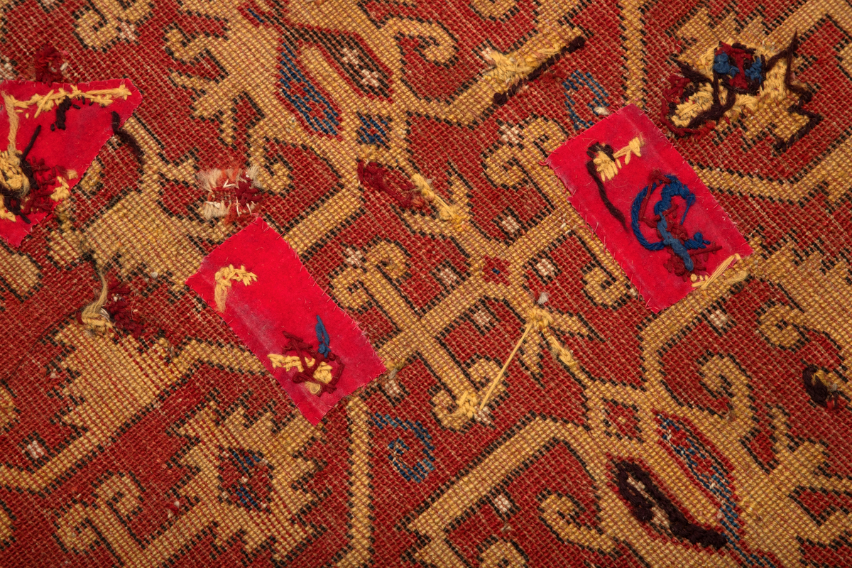 Håndlaget rektangulært teppe i ullfloss, utført med ghiordesknuter. Tett sammenhengende geometriske figurer i gult med litt blått og med svarte konturer på rød bunn. En klar blå bord rammer inn midtmotivet. Denne er dekorert med svarte og røde stiliserte blomster i ranke.