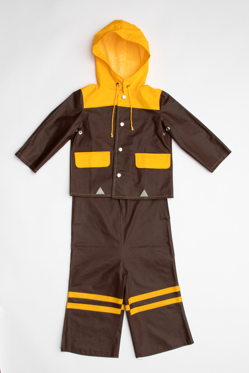 Regntøy for barn, cirka 2 år. Jakke og selebukse i maskinsydd og sveiset brunt og gult PVC-belagt bomullsstoff. Jakken er brun med rett fasong og påsveiset gul hette. Knappet foran med fire blanke trykknapper i hvitt metall. En lomme på hver side med gul klaff. Små, grå refleks-trekanter sveiset på langs nederkant. Buksen er knappet over skuldrene med trykknapper, kan knappes i tre høyder. Åpning i hver side med trykknapp. Langs ringningskantene påsydd gråhvite kantebånd. På hvert bukseben to gule, horisontale striper foran, en bak.