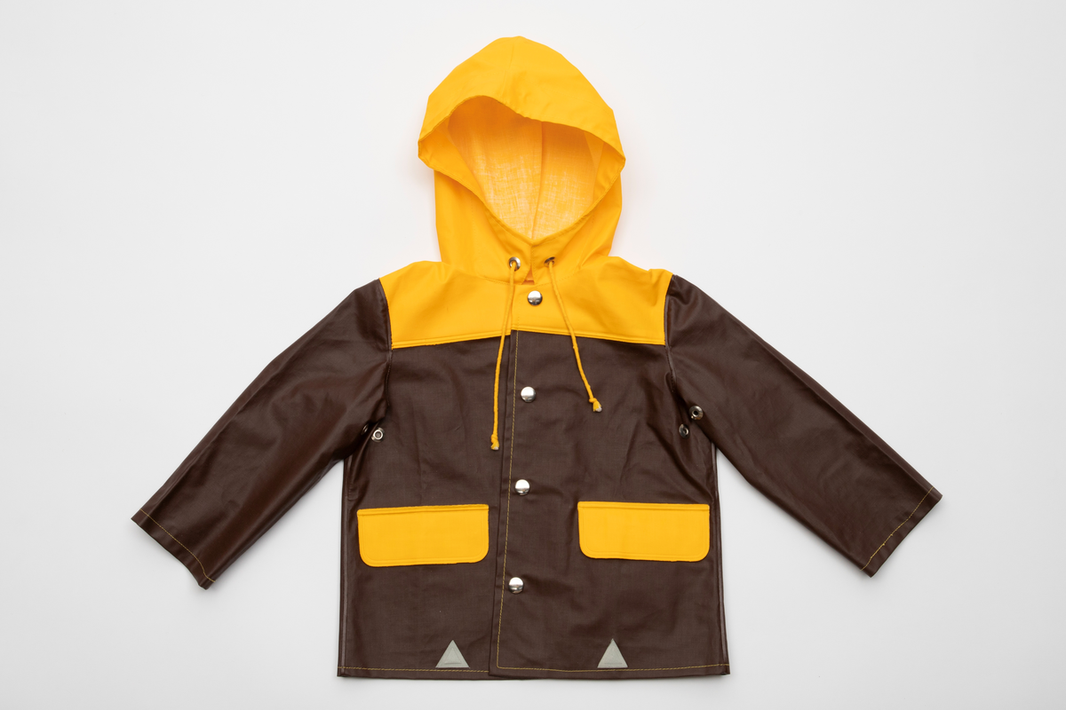 Regntøy for barn, cirka 2 år. Jakke og selebukse i maskinsydd og sveiset brunt og gult PVC-belagt bomullsstoff. Jakken er brun med rett fasong og påsveiset gul hette. Knappet foran med fire blanke trykknapper i hvitt metall. En lomme på hver side med gul klaff. Små, grå refleks-trekanter sveiset på langs nederkant. Buksen er knappet over skuldrene med trykknapper, kan knappes i tre høyder. Åpning i hver side med trykknapp. Langs ringningskantene påsydd gråhvite kantebånd. På hvert bukseben to gule, horisontale striper foran, en bak.