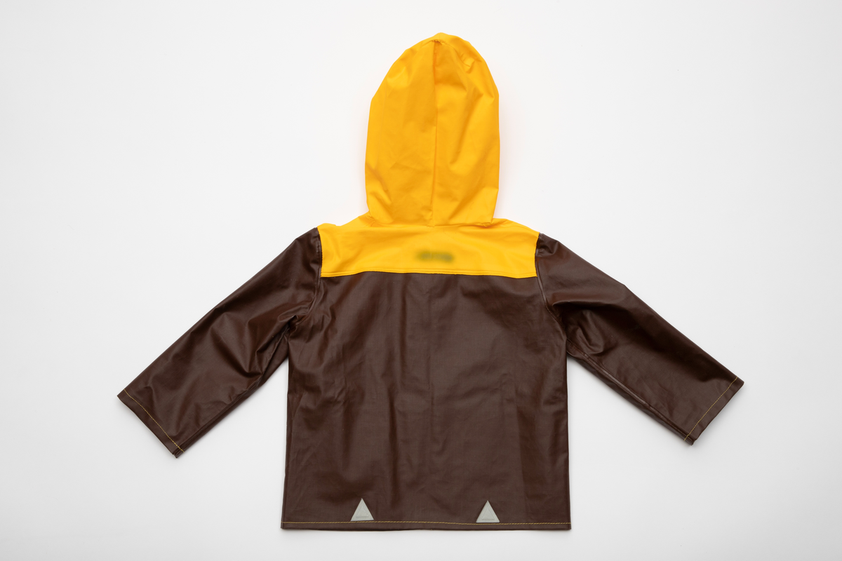 Regntøy for barn, cirka 2 år. Jakke og selebukse i maskinsydd og sveiset brunt og gult PVC-belagt bomullsstoff. Jakken er brun med rett fasong og påsveiset gul hette. Knappet foran med fire blanke trykknapper i hvitt metall. En lomme på hver side med gul klaff. Små, grå refleks-trekanter sveiset på langs nederkant. Buksen er knappet over skuldrene med trykknapper, kan knappes i tre høyder. Åpning i hver side med trykknapp. Langs ringningskantene påsydd gråhvite kantebånd. På hvert bukseben to gule, horisontale striper foran, en bak.
