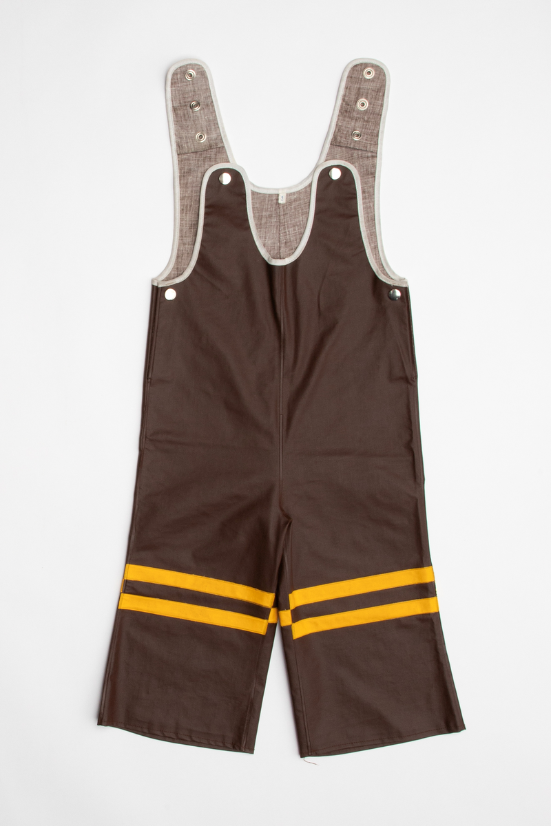 Regntøy for barn, cirka 2 år. Jakke og selebukse i maskinsydd og sveiset brunt og gult PVC-belagt bomullsstoff. Jakken er brun med rett fasong og påsveiset gul hette. Knappet foran med fire blanke trykknapper i hvitt metall. En lomme på hver side med gul klaff. Små, grå refleks-trekanter sveiset på langs nederkant. Buksen er knappet over skuldrene med trykknapper, kan knappes i tre høyder. Åpning i hver side med trykknapp. Langs ringningskantene påsydd gråhvite kantebånd. På hvert bukseben to gule, horisontale striper foran, en bak.