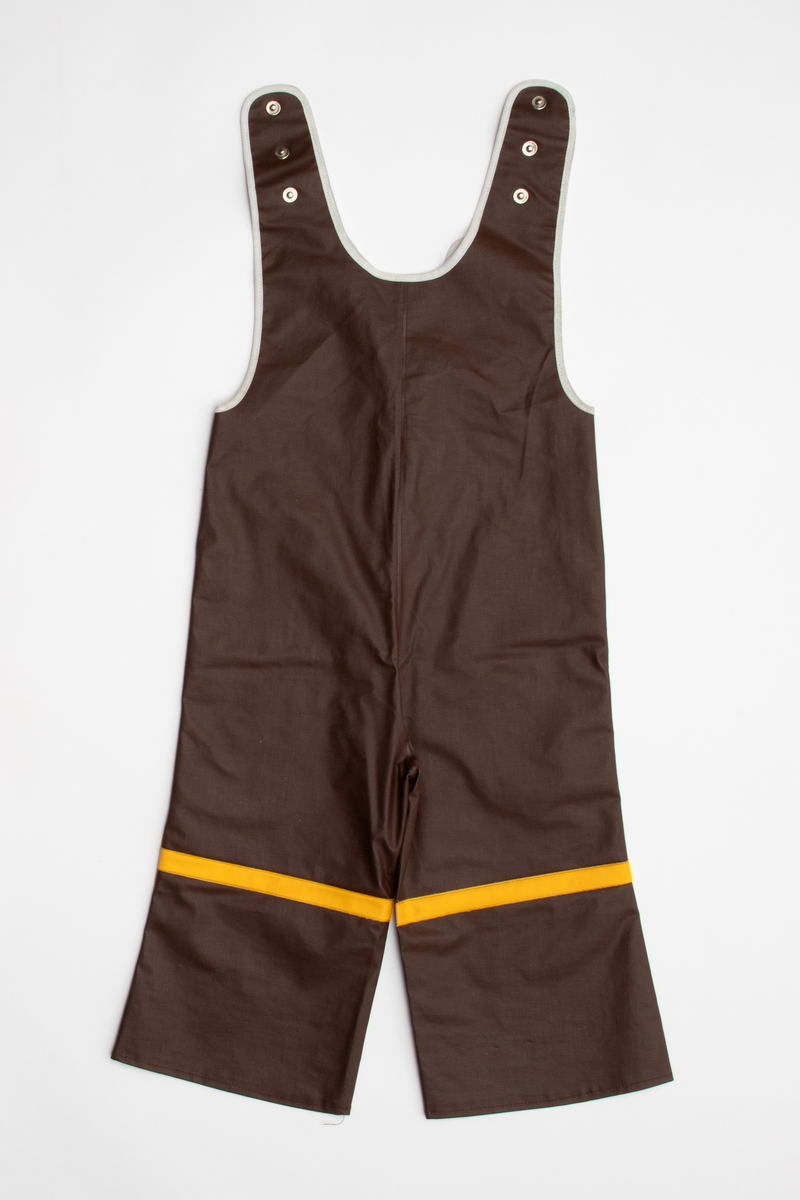 Regntøy for barn, cirka 2 år. Jakke og selebukse i maskinsydd og sveiset brunt og gult PVC-belagt bomullsstoff. Jakken er brun med rett fasong og påsveiset gul hette. Knappet foran med fire blanke trykknapper i hvitt metall. En lomme på hver side med gul klaff. Små, grå refleks-trekanter sveiset på langs nederkant. Buksen er knappet over skuldrene med trykknapper, kan knappes i tre høyder. Åpning i hver side med trykknapp. Langs ringningskantene påsydd gråhvite kantebånd. På hvert bukseben to gule, horisontale striper foran, en bak.