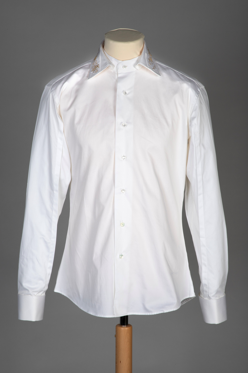 Hvit herreskjorte av bomullssateng. Betegnes som "hand grade", dvs. delvis håndarbeid selv om den er maskinsydd. Talllrike detaljer gjør skjorten meget spesiell: ettsøms ermer flankert av sydde legg og med doble, fôrede mansjetter isatt knuteformede mansjettknapper av hvit, elastisk, silkelignende snor. Innvendig knappestolpe à la smokinskjorte midt foran, men i motsetning til smokingskjorten, synes den ikke på retten. Skjorten lukkes med fem kuleformede, løse knapper, som har flat bakside og kan vendes. Den øverste sitter i halslinningen. De to nederste knappene er vanlige perlemorsskjorteknapper. Skjortens rygg har bærestykke og nedenfor to horisontale, sydde legg. På venstre side har den også et vertikalt "pyntelegg". Skjorten kan bæres med to ulike løssnipper og linningen har derfor fem små knapphull for feste av snipp. Alle vanlige sømmer er en slags indresøm.