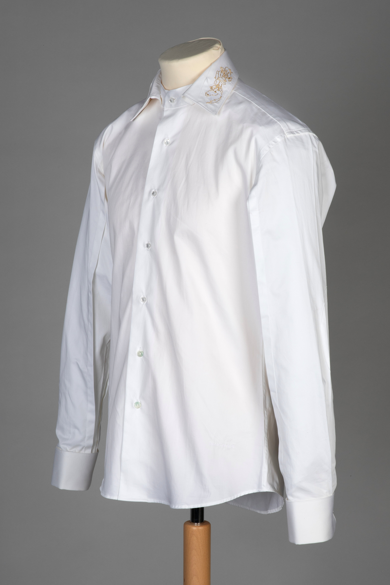 Hvit herreskjorte av bomullssateng. Betegnes som "hand grade", dvs. delvis håndarbeid selv om den er maskinsydd. Talllrike detaljer gjør skjorten meget spesiell: ettsøms ermer flankert av sydde legg og med doble, fôrede mansjetter isatt knuteformede mansjettknapper av hvit, elastisk, silkelignende snor. Innvendig knappestolpe à la smokinskjorte midt foran, men i motsetning til smokingskjorten, synes den ikke på retten. Skjorten lukkes med fem kuleformede, løse knapper, som har flat bakside og kan vendes. Den øverste sitter i halslinningen. De to nederste knappene er vanlige perlemorsskjorteknapper. Skjortens rygg har bærestykke og nedenfor to horisontale, sydde legg. På venstre side har den også et vertikalt "pyntelegg". Skjorten kan bæres med to ulike løssnipper og linningen har derfor fem små knapphull for feste av snipp. Alle vanlige sømmer er en slags indresøm.
