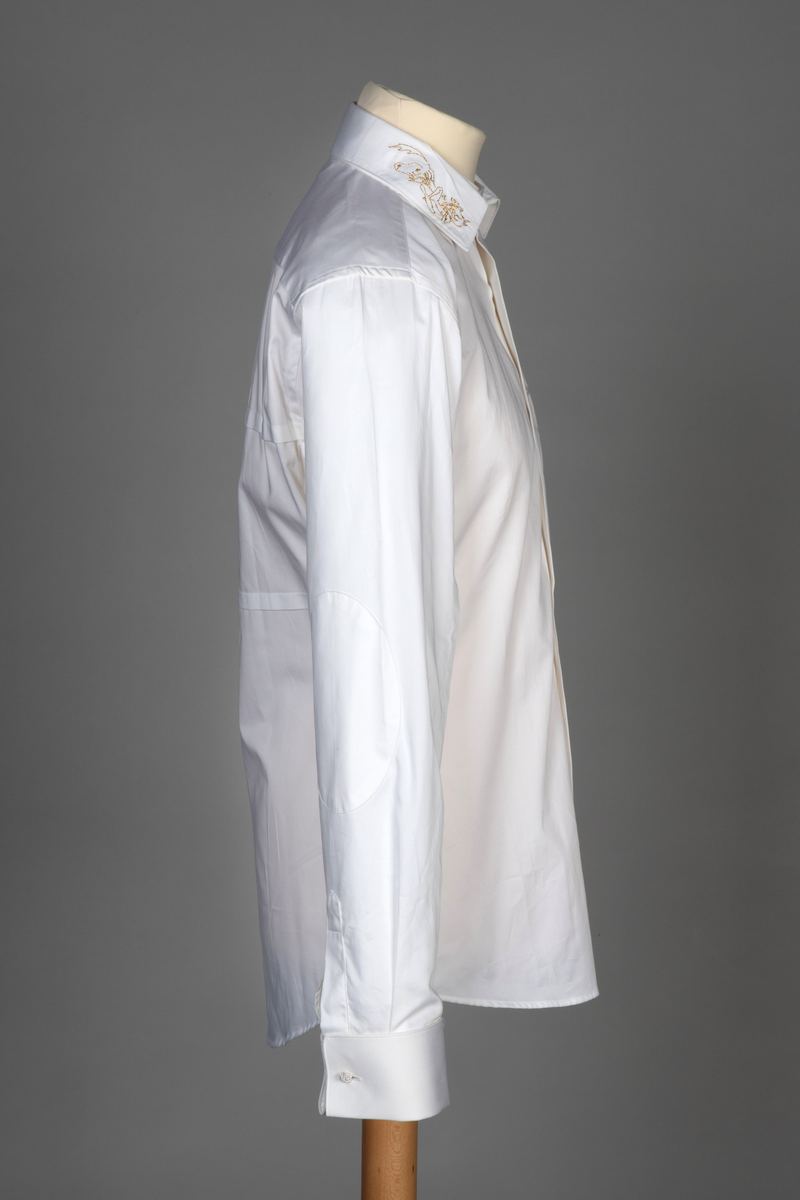 Hvit herreskjorte av bomullssateng. Betegnes som "hand grade", dvs. delvis håndarbeid selv om den er maskinsydd. Talllrike detaljer gjør skjorten meget spesiell: ettsøms ermer flankert av sydde legg og med doble, fôrede mansjetter isatt knuteformede mansjettknapper av hvit, elastisk, silkelignende snor. Innvendig knappestolpe à la smokinskjorte midt foran, men i motsetning til smokingskjorten, synes den ikke på retten. Skjorten lukkes med fem kuleformede, løse knapper, som har flat bakside og kan vendes. Den øverste sitter i halslinningen. De to nederste knappene er vanlige perlemorsskjorteknapper. Skjortens rygg har bærestykke og nedenfor to horisontale, sydde legg. På venstre side har den også et vertikalt "pyntelegg". Skjorten kan bæres med to ulike løssnipper og linningen har derfor fem små knapphull for feste av snipp. Alle vanlige sømmer er en slags indresøm.