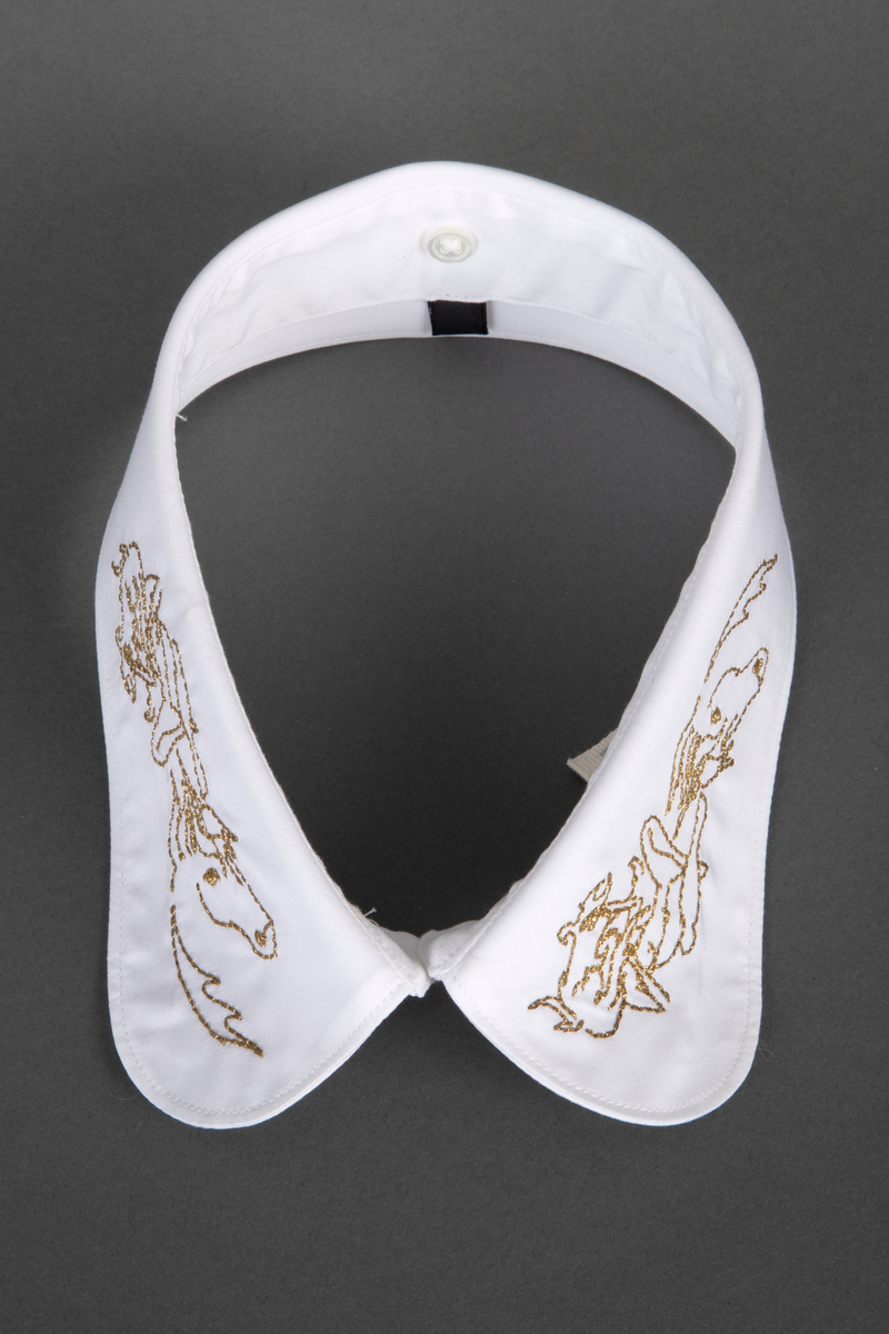 Løssnipp, innvendig fôret med avrundede snipphjørner. Forsiden er prydet på begge snipper med kjedestingsbroderi i gulltråd, mens baksiden er ensfarget hvit. Broderiet forestiller et enhjørningshode under en sittende kvinnefigur, som ser ut til å spille en fele. På kragestanden er påsydd fem små knapper til å kneppe på skjortens halslinning.