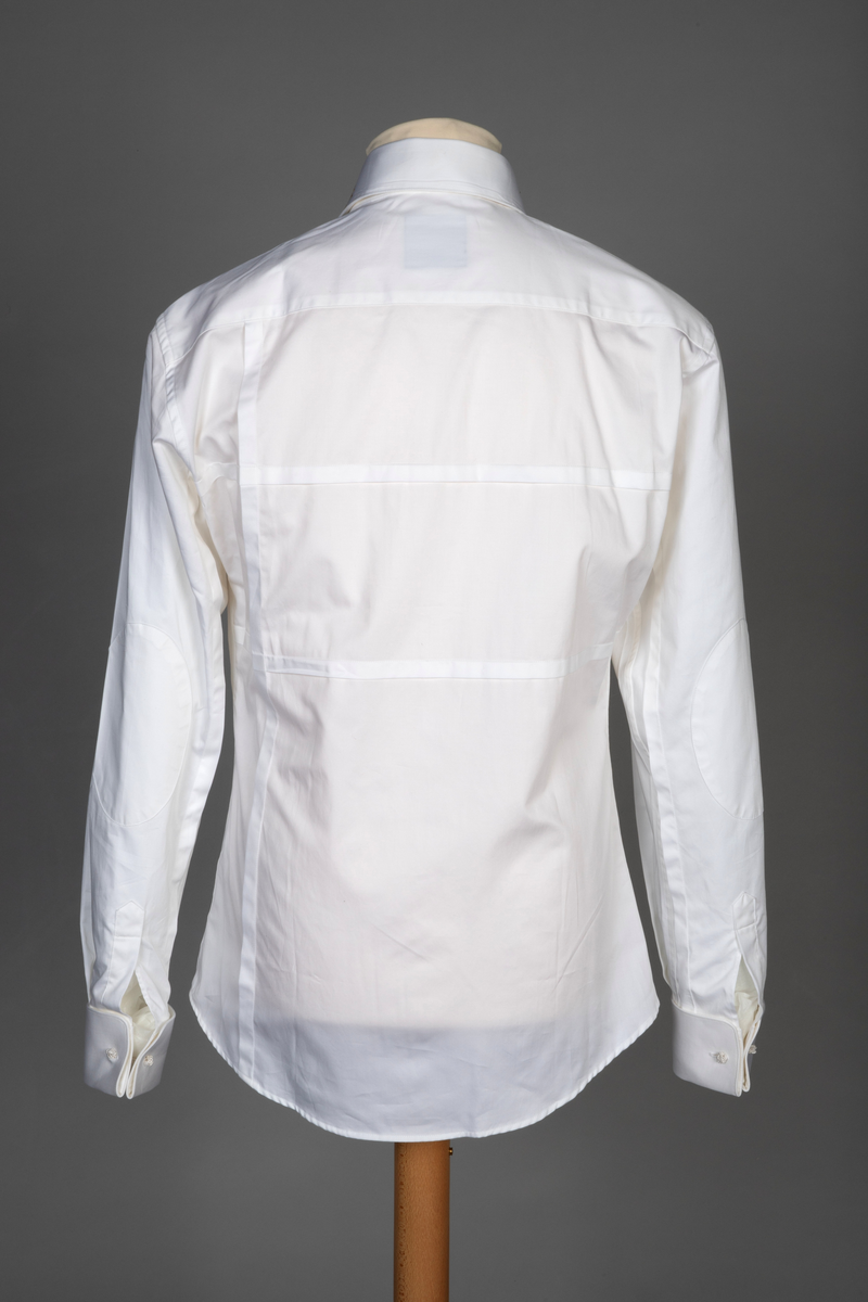 Hvit herreskjorte av bomullssateng. Betegnes som "hand grade", dvs. delvis håndarbeid selv om den er maskinsydd. Talllrike detaljer gjør skjorten meget spesiell: ettsøms ermer flankert av sydde legg og med doble, fôrede mansjetter isatt knuteformede mansjettknapper av hvit, elastisk, silkelignende snor. Innvendig knappestolpe à la smokinskjorte midt foran, men i motsetning til smokingskjorten, synes den ikke på retten. Skjorten lukkes med fem kuleformede, løse knapper, som har flat bakside og kan vendes. Den øverste sitter i halslinningen. De to nederste knappene er vanlige perlemorsskjorteknapper. Skjortens rygg har bærestykke og nedenfor to horisontale, sydde legg. På venstre side har den også et vertikalt "pyntelegg". Skjorten kan bæres med to ulike løssnipper og linningen har derfor fem små knapphull for feste av snipp. Alle vanlige sømmer er en slags indresøm.