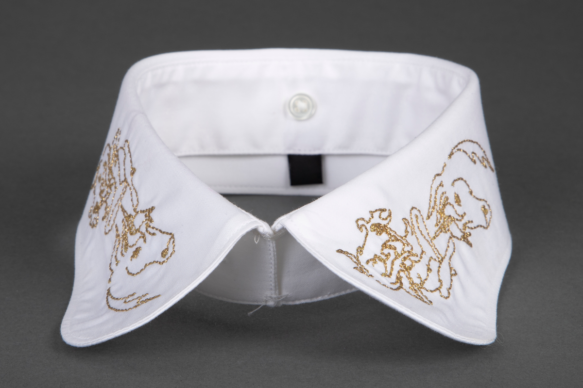 Løssnipp, innvendig fôret med avrundede snipphjørner. Forsiden er prydet på begge snipper med kjedestingsbroderi i gulltråd, mens baksiden er ensfarget hvit. Broderiet forestiller et enhjørningshode under en sittende kvinnefigur, som ser ut til å spille en fele. På kragestanden er påsydd fem små knapper til å kneppe på skjortens halslinning.