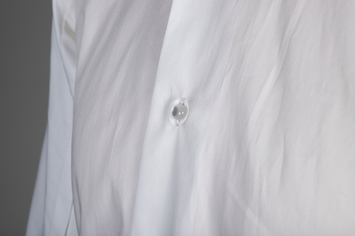 Hvit herreskjorte av bomullssateng. Betegnes som "hand grade", dvs. delvis håndarbeid selv om den er maskinsydd. Talllrike detaljer gjør skjorten meget spesiell: ettsøms ermer flankert av sydde legg og med doble, fôrede mansjetter isatt knuteformede mansjettknapper av hvit, elastisk, silkelignende snor. Innvendig knappestolpe à la smokinskjorte midt foran, men i motsetning til smokingskjorten, synes den ikke på retten. Skjorten lukkes med fem kuleformede, løse knapper, som har flat bakside og kan vendes. Den øverste sitter i halslinningen. De to nederste knappene er vanlige perlemorsskjorteknapper. Skjortens rygg har bærestykke og nedenfor to horisontale, sydde legg. På venstre side har den også et vertikalt "pyntelegg". Skjorten kan bæres med to ulike løssnipper og linningen har derfor fem små knapphull for feste av snipp. Alle vanlige sømmer er en slags indresøm.