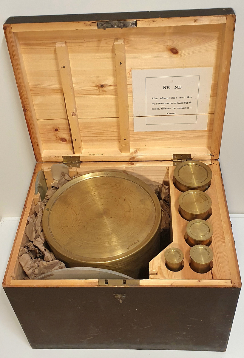 Stor kasse av tre med plass til seks hulmål i messing og tre glassplater. 

B: 20L
C: 5L
D: 1L
E: 0,5L
F: 0,2L
G: 0,1LL
H: 0,05L