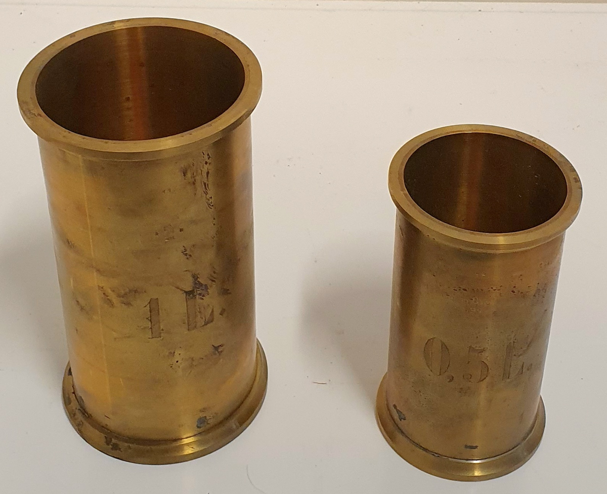 Stor kasse av tre med plass til seks hulmål i messing og tre glassplater. 

B: 20L
C: 5L
D: 1L
E: 0,5L
F: 0,2L
G: 0,1LL
H: 0,05L