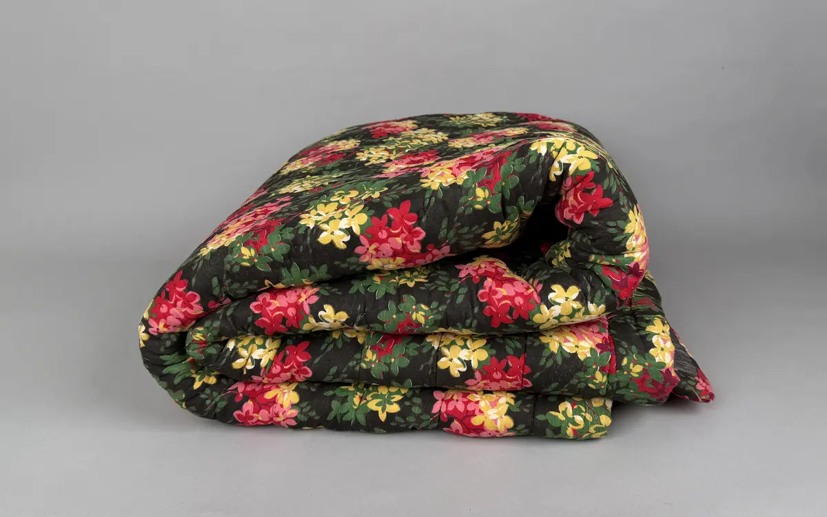 Ett enkelt stoppat vaddtäcke med ovansida av blommigt mönstrat bomullstyg, i rött och gult på mörk botten. Baksida av (troligen) halvlinne i svart/vit tuskaft.
Täcket är oanvänt, något skört på ovansidan.
