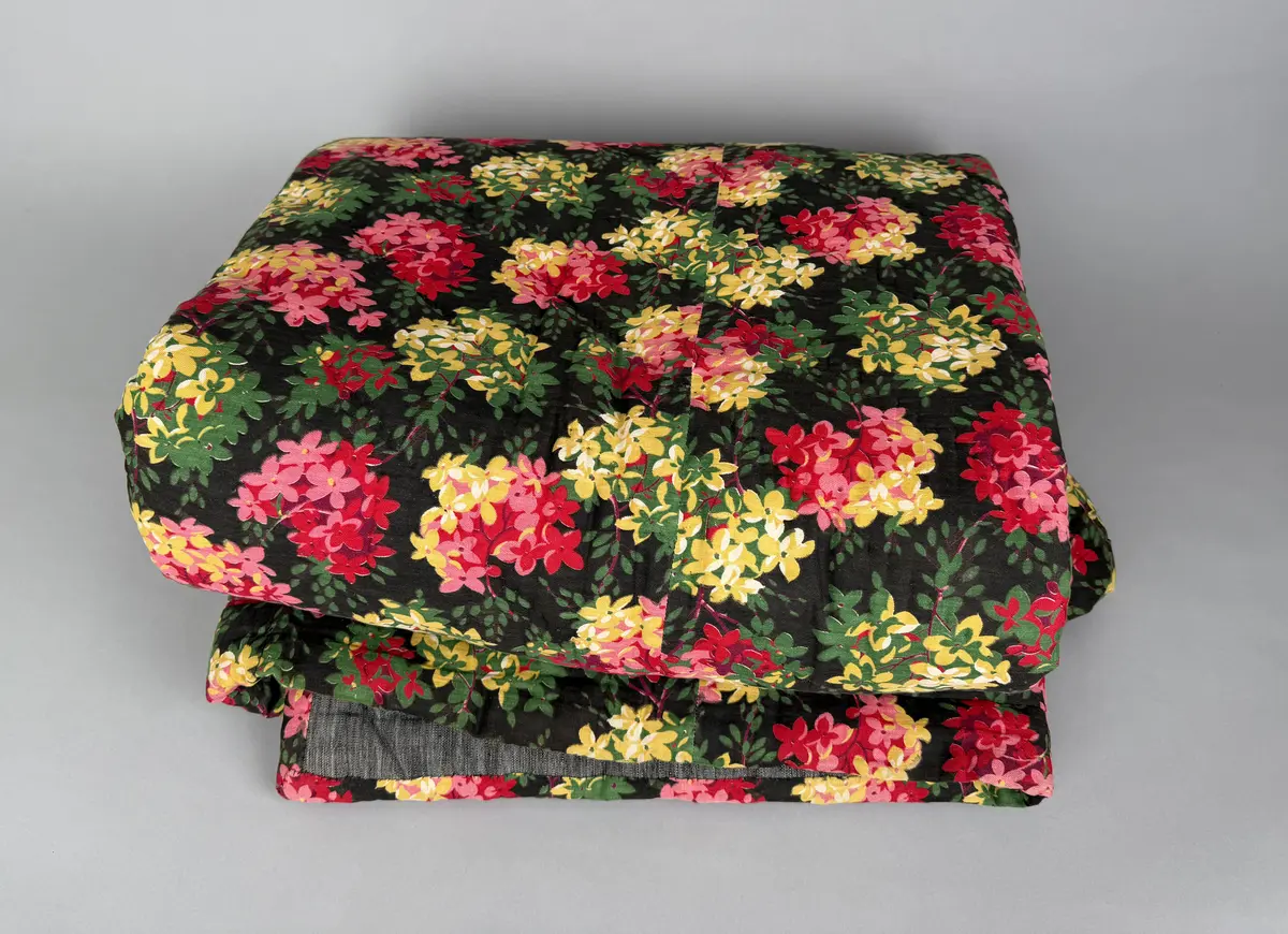 Ett enkelt stoppat vaddtäcke med ovansida av blommigt mönstrat bomullstyg, i rött och gult på mörk botten. Baksida av (troligen) halvlinne i svart/vit tuskaft.
Täcket är oanvänt, något skört på ovansidan.
