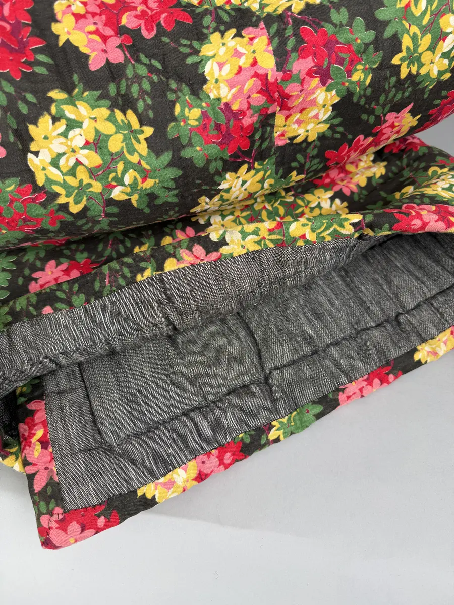 Ett enkelt stoppat vaddtäcke med ovansida av blommigt mönstrat bomullstyg, i rött och gult på mörk botten. Baksida av (troligen) halvlinne i svart/vit tuskaft.
Täcket är oanvänt, något skört på ovansidan.
