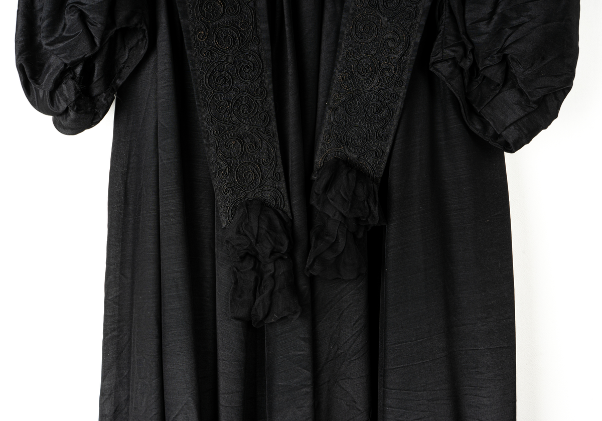 a, Klädning av svart alpackasiden. Prydd med bårder i svart.
b, garnering till livet.
c, kjol med bård och veck som pryder kanten.