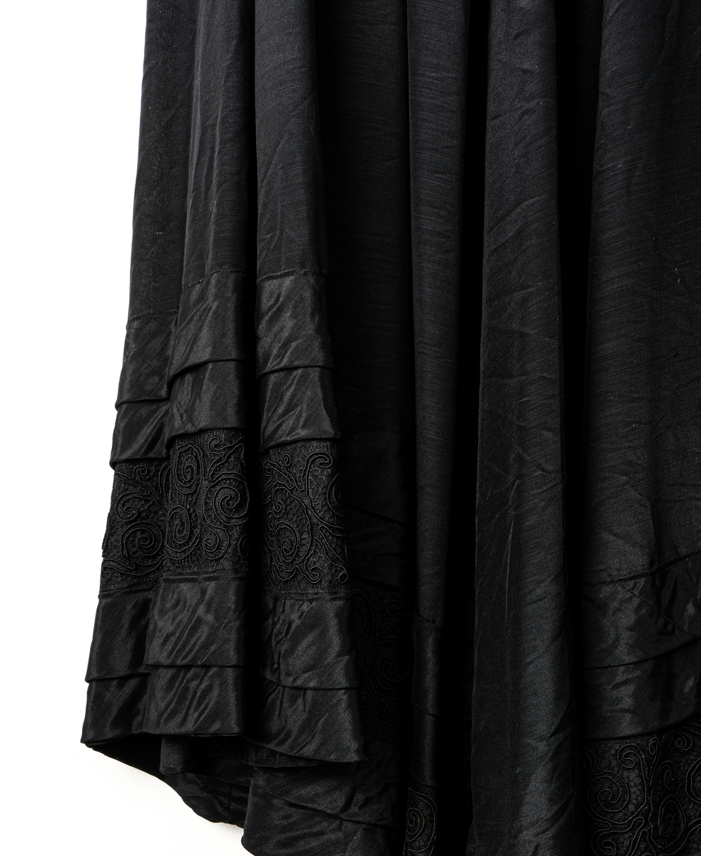 a, Klädning av svart alpackasiden. Prydd med bårder i svart.
b, garnering till livet.
c, kjol med bård och veck som pryder kanten.