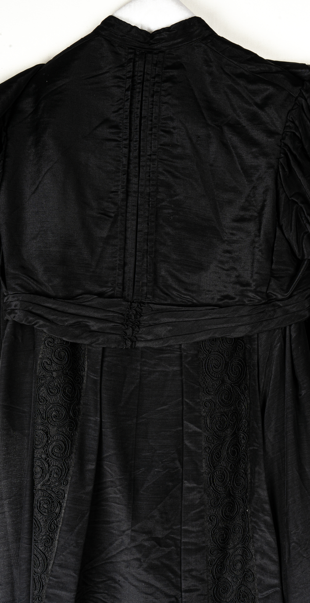 a, Klädning av svart alpackasiden. Prydd med bårder i svart.
b, garnering till livet.
c, kjol med bård och veck som pryder kanten.