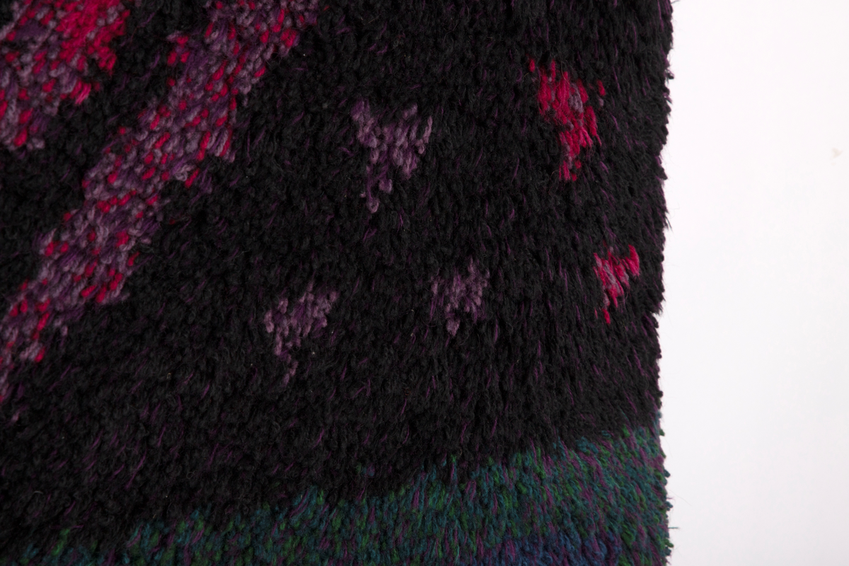 Rektangulær rye i ull med grenformet motiv bestående av striper i svart, rosa og fiolett. De horisontale partiene oppe og nede er sjattert i grønt, lilla, fiolett og blått.