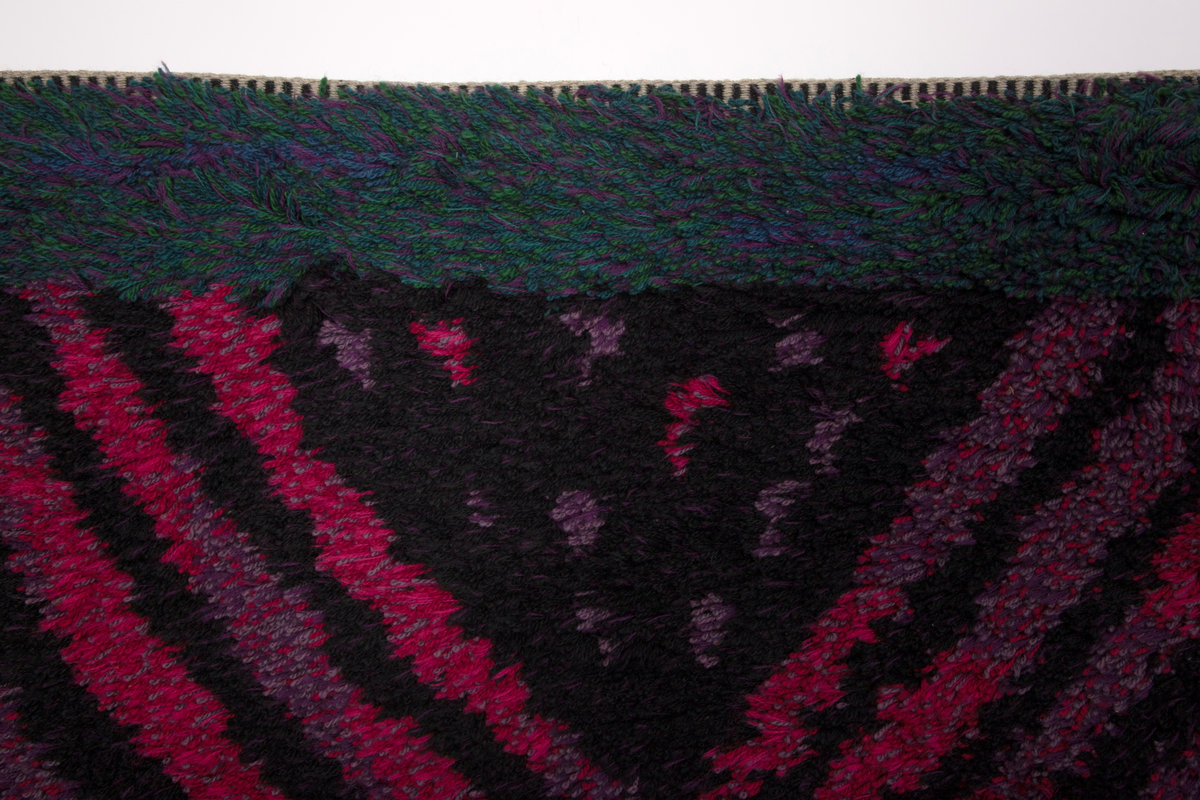 Rektangulær rye i ull med grenformet motiv bestående av striper i svart, rosa og fiolett. De horisontale partiene oppe og nede er sjattert i grønt, lilla, fiolett og blått.