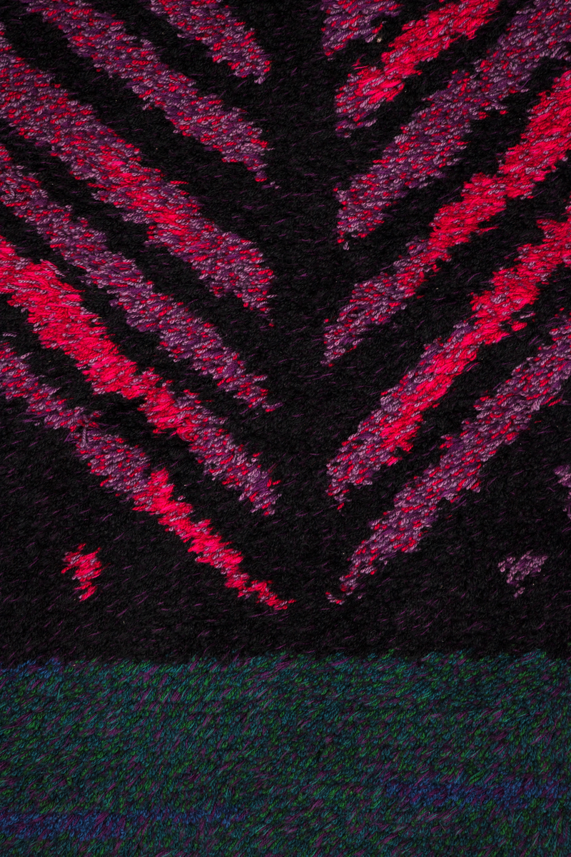 Rektangulær rye i ull med grenformet motiv bestående av striper i svart, rosa og fiolett. De horisontale partiene oppe og nede er sjattert i grønt, lilla, fiolett og blått.