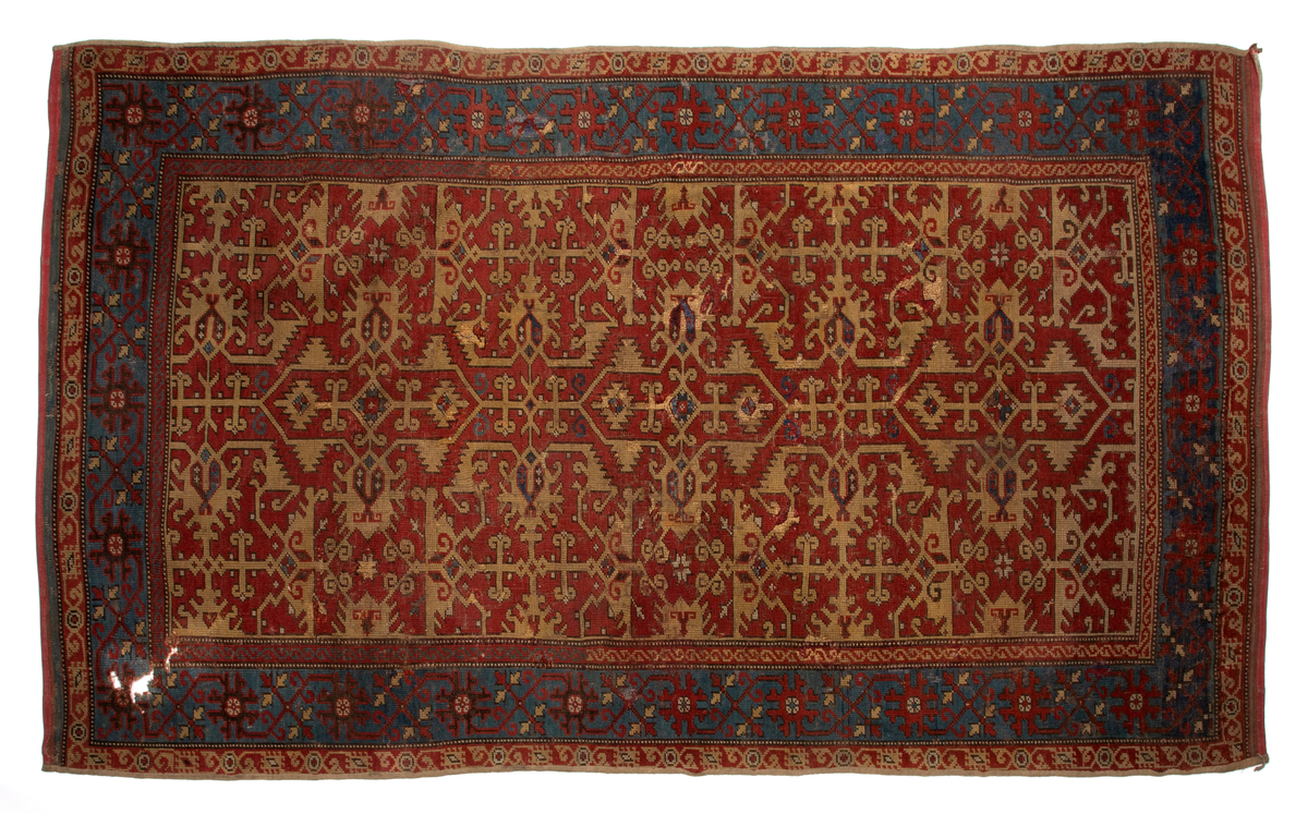 Håndlaget rektangulært teppe i ullfloss, utført med ghiordesknuter. Tett sammenhengende geometriske figurer i gult med litt blått og med svarte konturer på rød bunn. En klar blå bord rammer inn midtmotivet. Denne er dekorert med svarte og røde stiliserte blomster i ranke.