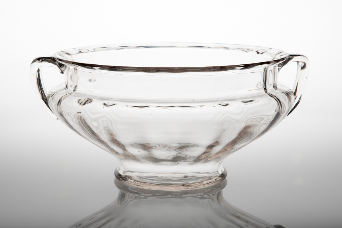 Skål i klart glass. Rund, buket korpus på lav fot med vertikalt riflede innervegger. To c-formede håndtak diametralt påsatte på korpus.