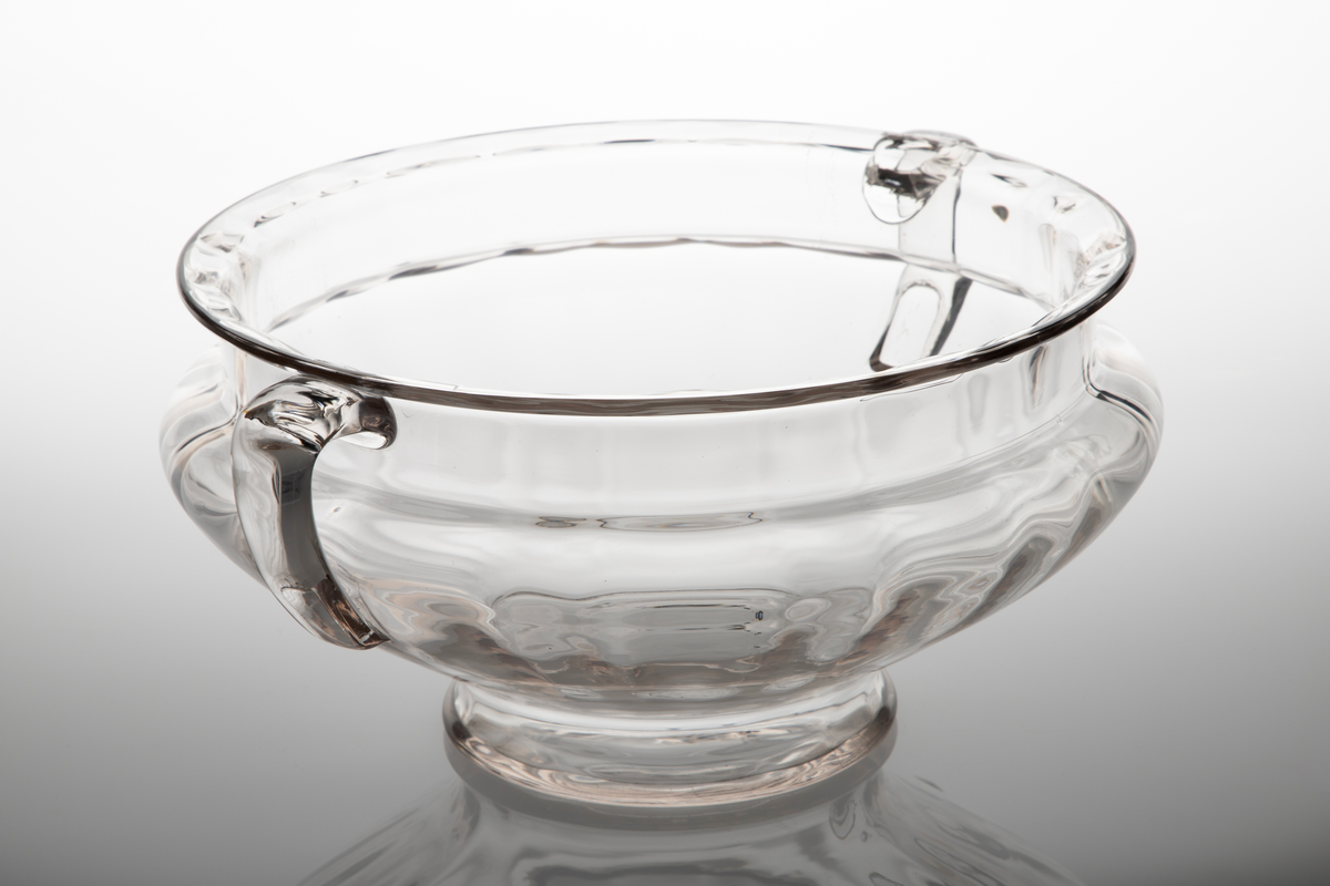 Skål i klart glass. Rund, buket korpus på lav fot med vertikalt riflede innervegger. To c-formede håndtak diametralt påsatte på korpus.