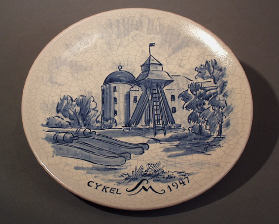 Tallrik av keramik med vit halvmatt, krakelerad glasyr och handmålat motiv i blått: Gunilla-klockan med Uppsala slott i bakgrunden. Text: Cykel SM 1947. På baksidan; U.C.K. - FYRISHOF - UNI. Ståltråd för upphängning.