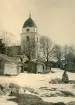Bild av Rättviks kyrka i Dalarna samt kyrkstallarna i förgrunden, en vintrig dag 1926.