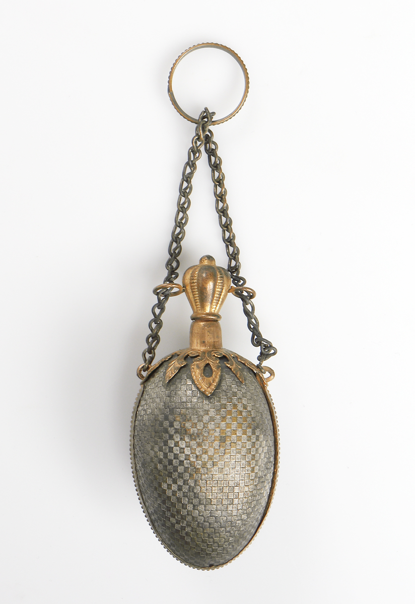 Äggformad luktflaska av grå metall med rutmönster i relief. Detaljer samt kork av gul metall. Upptill en liten kedja med ring, även dessa i gul metall. För förvaring av luktsalt. 

Inskrivet i huvudbok år 1969.