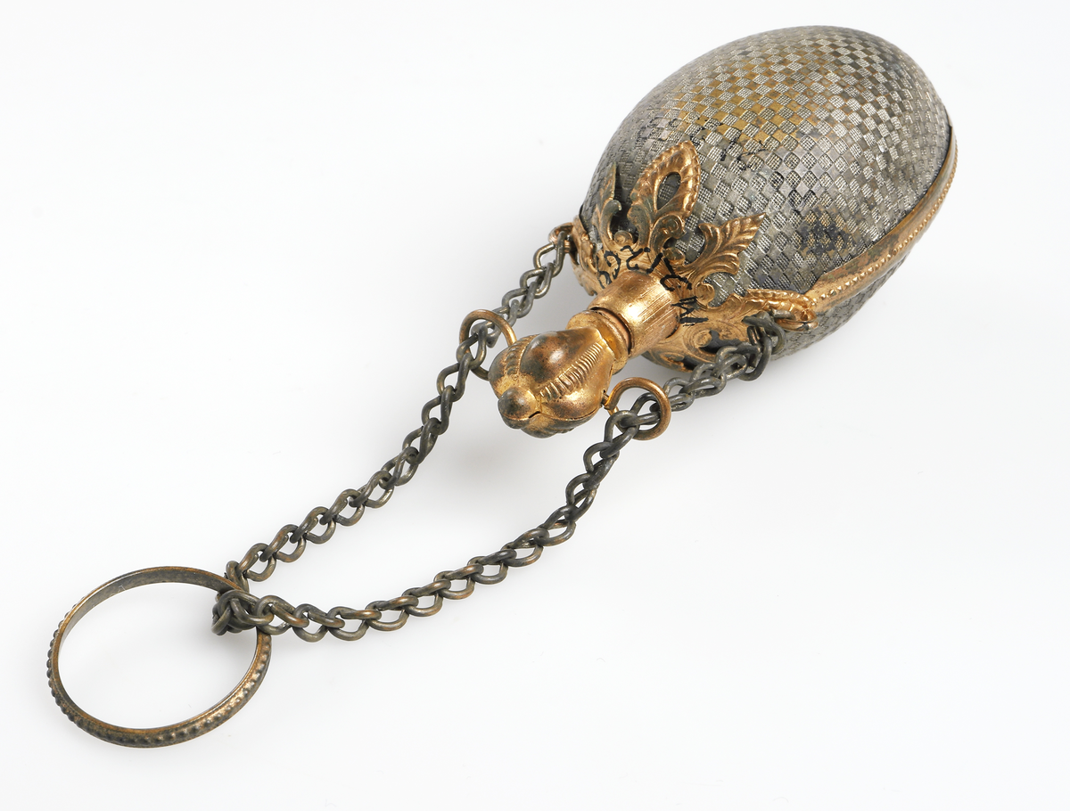 Äggformad luktflaska av grå metall med rutmönster i relief. Detaljer samt kork av gul metall. Upptill en liten kedja med ring, även dessa i gul metall. För förvaring av luktsalt. 

Inskrivet i huvudbok år 1969.