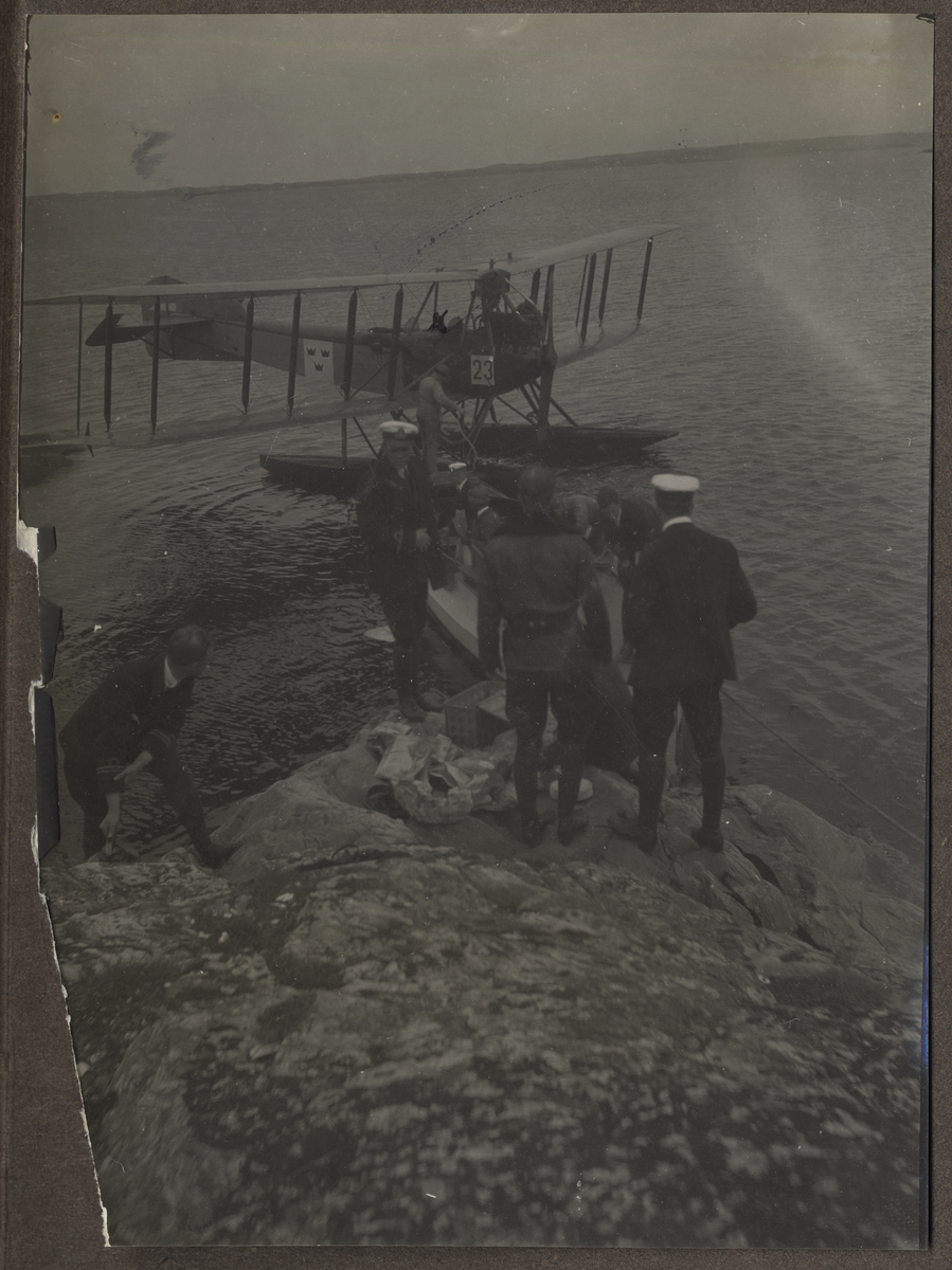 Bilden visar en grupp officerar från Marinflyget som har packat ihop efter lunchen på en klippa i skärgården och håller på att lasta flygplanet Friedrichshafen FF 49 i bakgrunden.