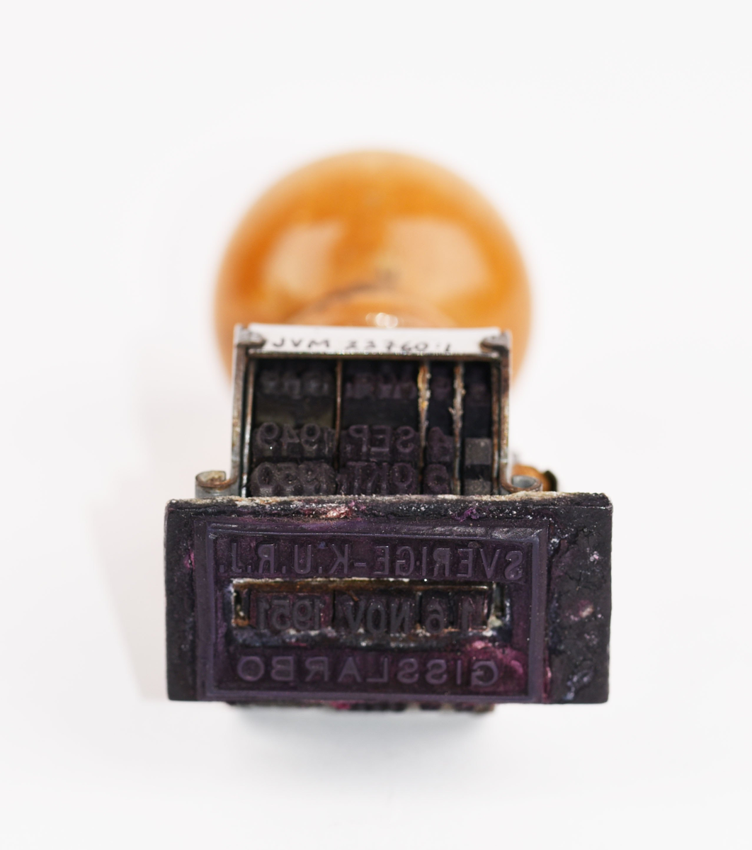 Datumstämpel (:1) i tillhörande pappask (:2) med lock (:3). Datumstämpeln har texten "SVERIGE - K.U.R.J. XX MÅN ÅRTAL GISSLARBO" innanför en rektangulär kantlinje. Den är framskruvad till "16 NOV. 1951". Stämpeln har en rund knopp av ljust svarvat lackat trä med en mässingsnit som visar vilken sida som ska vara vänd mot brukaren för rättvänd stämpling. På askens insida (:2) finns några stämpelavtryck i olika kulörer. På locket (:3) är det stämplat "SVERIGE - K.U.R.J. 22 NOV 1951" samt ifyllt "GISSLARBO". På locket (:3) står det stämplat "SWEDISH", sedan fortsätter texten på askens underdel (:2) "BRASS" samt "No. 2 All Square Dater Plate 3".