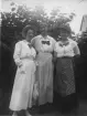 Tre kvinnor avbildade stående utomhus framför en lövklädd buske. Ljusa blusar med mörka rosetter vid kragen bärs av samtliga. Lång kjol respektive klänning samt olika huvudbonader förekommer. En byggnad skymtar i bakgrunden. Tidigt 1900-tal.