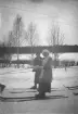 Två personer avbildade stående på skidor i snötäckt landskap. Vinterklädsel med kappor och huvudbonader förekommer. I bakgrunden ses lövträd, buskage och skogsbryn samt överliggande ledningar. Tidigt 1900-tal.