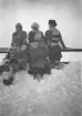 En grupp människor avbildade sittande på en snövall utomhus. Vinterklädsel med kappor, mössor, halsdukar och vantar förekommer. I bakgrunden ses ett räcke samt öppet snötäckt landskap. Tidigt 1900-tal.